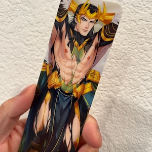 May include: A rectangular bookmark featuring a stylized illustration of a character with long black hair, a gold crown, and green and gold attire. The character is depicted with their arms behind their head, and the bookmark has a white background.