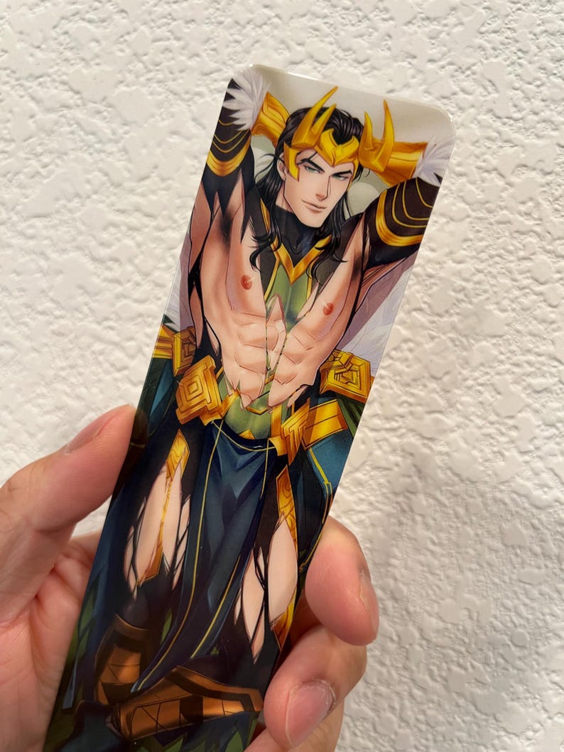May include: A rectangular bookmark featuring a stylized illustration of a character with long black hair, a gold crown, and green and gold attire. The character is depicted with their arms behind their head, and the bookmark has a white background.