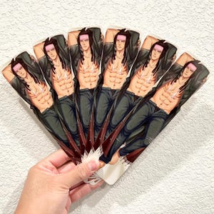 May include: A hand fan featuring a repeating illustration of a shirtless person in a leather jacket and jeans. The person has long dark hair, a pink headband, and is posed with their arms behind their head. The fan is held in a hand.