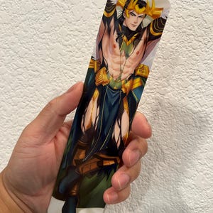 May include: A rectangular bookmark featuring a stylized illustration of a character with long black hair, a golden crown, and a green and gold outfit. The character is depicted with their arms behind their head.
