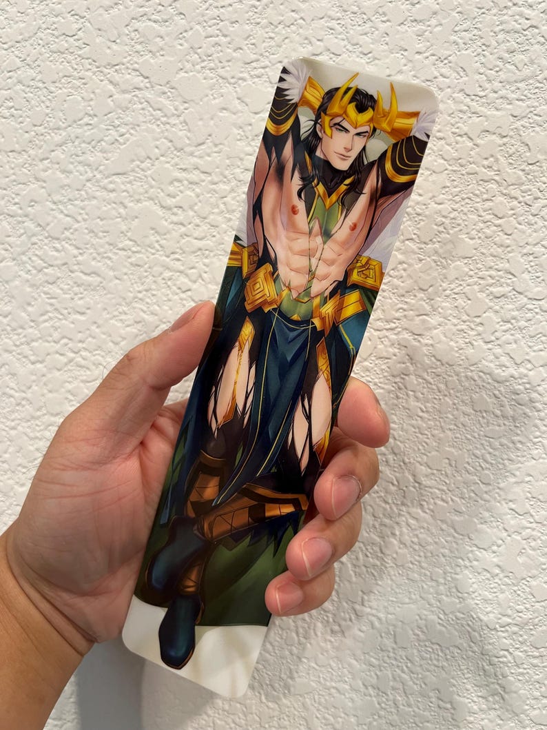 May include: A rectangular bookmark featuring a stylized illustration of a character with long black hair, a golden crown, and a green and gold outfit. The character is depicted with their arms behind their head.