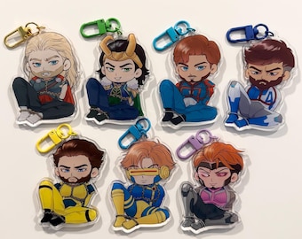 May include: A collection of seven acrylic character keychains. Each keychain features a cartoon-style illustration of a superhero or comic book character in a seated pose. The keychains have different colored clasps and are on a white background.