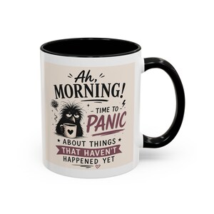 Peut inclure: Un mug en céramique blanche avec une anse et un bord intérieur noirs. L'avant présente le dessin d'une créature noire ébouriffée tenant une petite tasse, avec le texte : Ah, MORNING! TIME TO PANIC ABOUT THINGS THAT HAVEN'T HAPPENED YET.