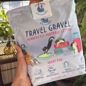 Op de afbeelding: Een zak "Roaming Whiskers" reis kattenbakvulling. De zak is wit met een kleurrijke afbeelding van katten en bagage. De tekst luidt "Travel Gravel, Purrfectly Portable Litter". Nettogewicht: 1,8 kg.