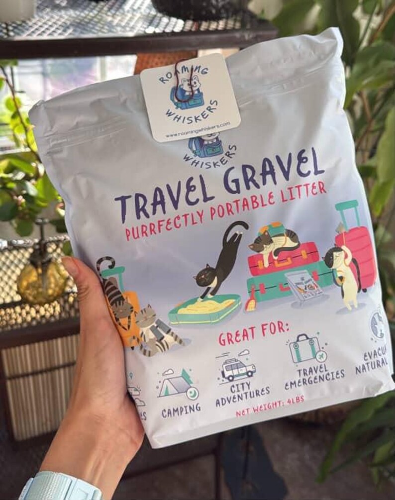 Op de afbeelding: Een zak "Roaming Whiskers" reis kattenbakvulling. De zak is wit met een kleurrijke afbeelding van katten en bagage. De tekst luidt "Travel Gravel, Purrfectly Portable Litter". Nettogewicht: 1,8 kg.