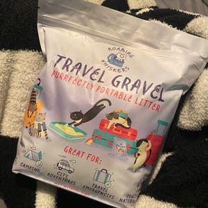 Op de afbeelding: Een zak "Roaming Whiskers Travel Gravel" kattenbakvulling. De zak is wit met illustraties van katten en bagage. Tekst op de zak luidt "Purrfectly Portable Litter" en "Great For: Camping, City Adventures, Travel Emergencies, Natural".