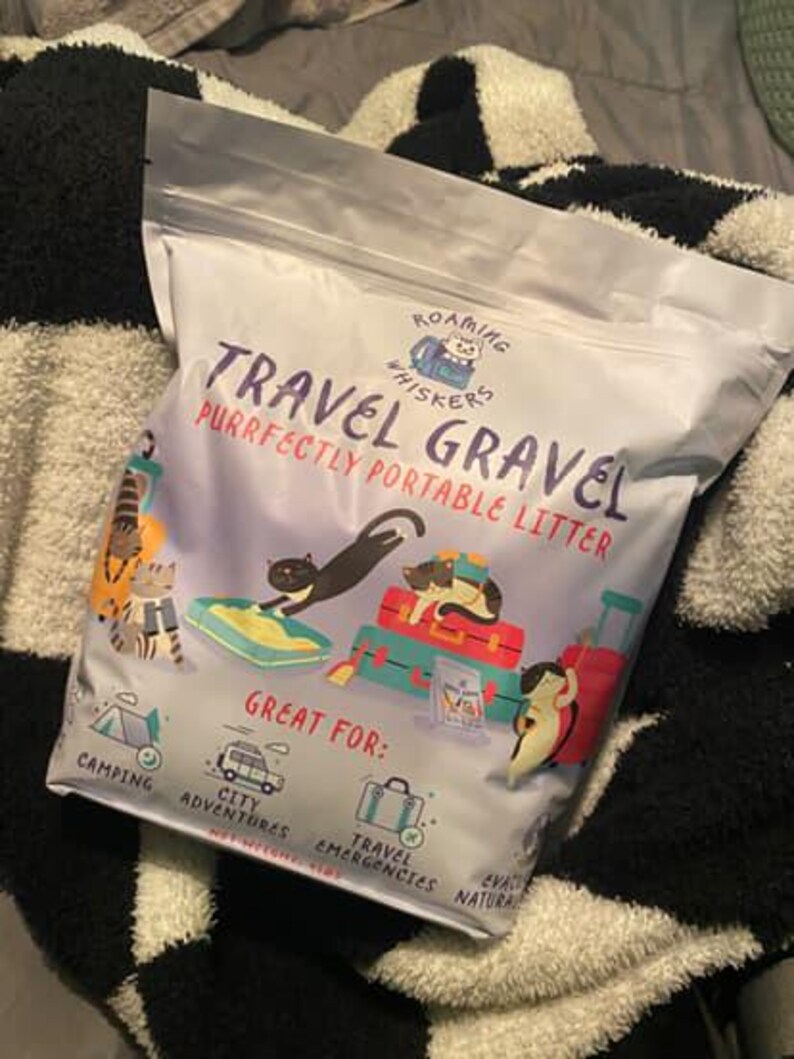 Op de afbeelding: Een zak "Roaming Whiskers Travel Gravel" kattenbakvulling. De zak is wit met illustraties van katten en bagage. Tekst op de zak luidt "Purrfectly Portable Litter" en "Great For: Camping, City Adventures, Travel Emergencies, Natural".
