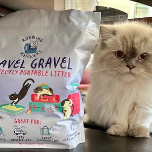 Op de afbeelding: Een zak "Roaming Whiskers Travel Gravel" kattenbakvulling naast een pluizige, lichtgekleurde kat. De zak bevat illustraties van katten en koffers, met tekst die aangeeft dat het "Purrfectly Portable Litter" is voor op reis.
