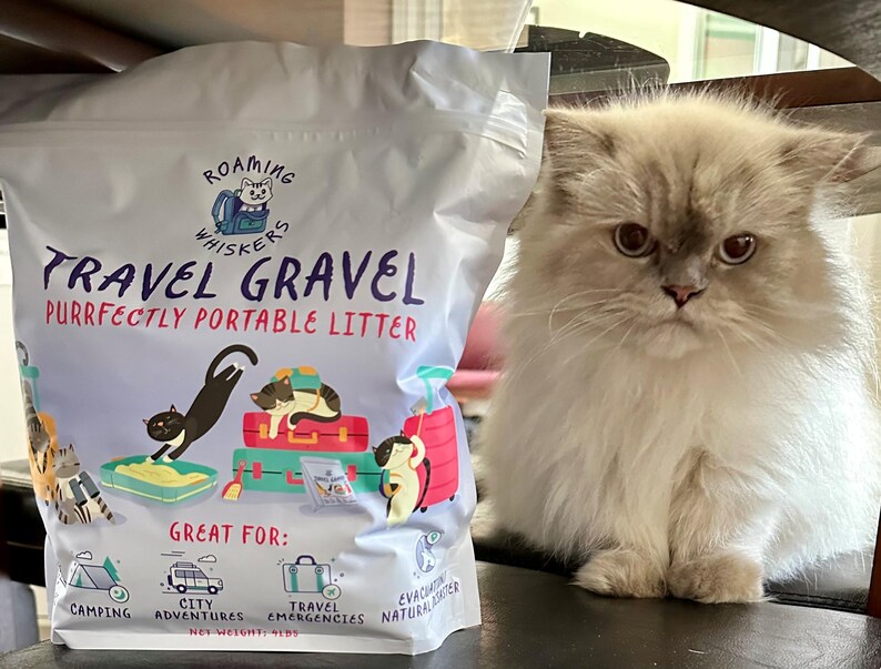Op de afbeelding: Een zak "Roaming Whiskers Travel Gravel" kattenbakvulling naast een pluizige, lichtgekleurde kat. De zak bevat illustraties van katten en koffers, met tekst die aangeeft dat het "Purrfectly Portable Litter" is voor op reis.