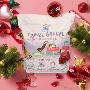 Op de afbeelding: Een witte zak "Travel Gravel" kattenbakvulling met illustraties van katten en bagage. De zak is omgeven door kerstversieringen en groen. De tekst op de zak luidt "Purrfectly Portable Litter" en "Great for: Camping, City Adventures, Travel Emergencies, Nature".