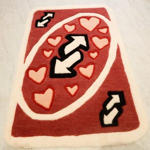 May include: A red and white rug shaped like an Uno reverse card. The rug features a large black and white reverse arrow symbol, surrounded by pink hearts. The edges are trimmed with a white border.