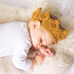 K&ouml;nnte beinhalten: Ein Baby mit einer gestrickten gelben Krone schl&auml;ft auf einer wei&szlig;en, flauschigen Decke.