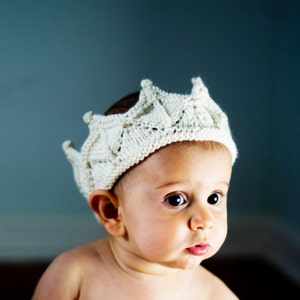 K&ouml;nnte beinhalten: Ein Baby tr&auml;gt eine wei&szlig;e Strickkrone. Die Krone hat ein detailliertes Design und ist aus weichem Garn gefertigt.