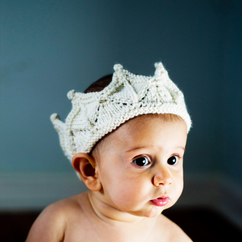 K&ouml;nnte beinhalten: Ein Baby tr&auml;gt eine wei&szlig;e Strickkrone. Die Krone hat ein detailliertes Design und ist aus weichem Garn gefertigt.