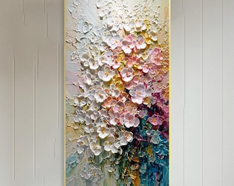 May include: A vertical floral painting with a gold frame. The artwork features a textured, three-dimensional effect with white, pink, yellow, and blue flowers. The painting is displayed above a white sofa in a well-lit living room.