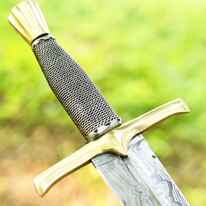 May include: Close-up of a sword with a gold-colored hilt and crossguard. The handle is wrapped in a silver-colored mesh. The blade has a patterned design. The sword is set against a blurred green background.