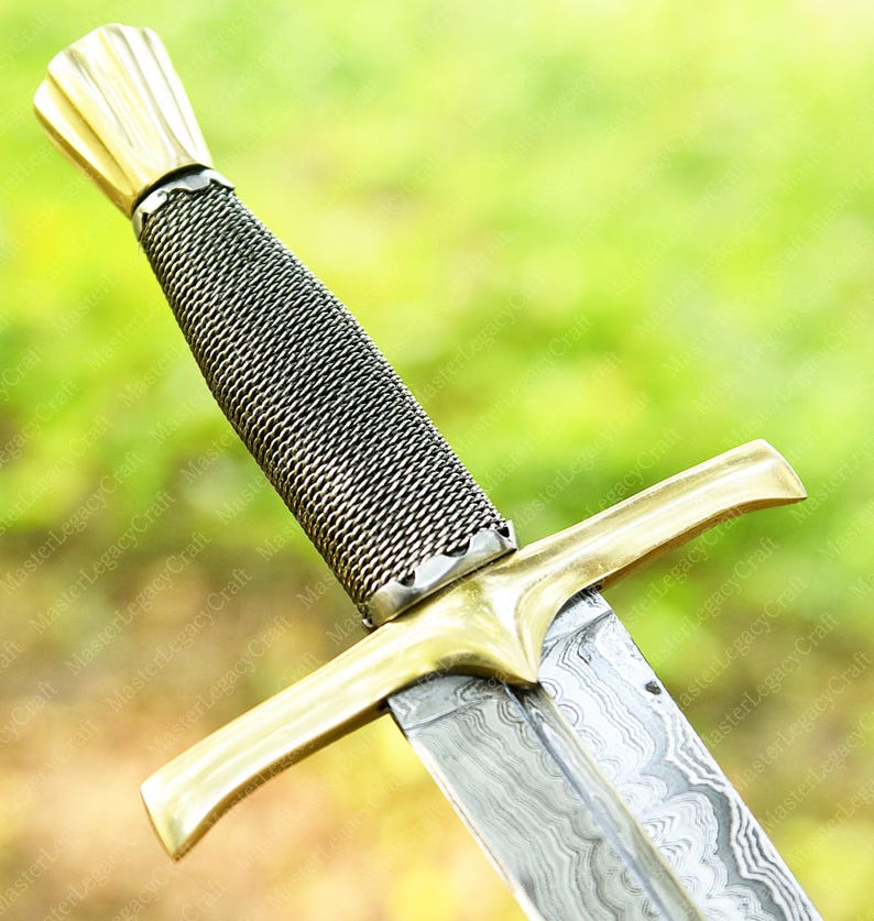 May include: Close-up of a sword with a gold-colored hilt and crossguard. The handle is wrapped in a silver-colored mesh. The blade has a patterned design. The sword is set against a blurred green background.