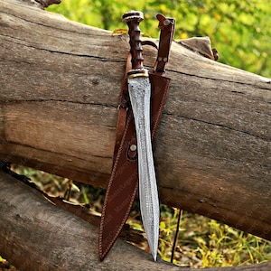 May include: A detailed image of a Roman Gladius sword with a brown wooden handle and a silver blade. The sword is sheathed in a brown leather scabbard. The sword and scabbard are resting on a large, weathered log.