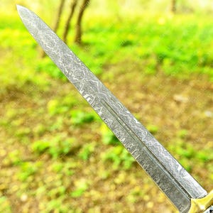 May include: Close-up of a decorative sword blade with a detailed, patterned design. The blade has a sharp point and a central groove. The handle is not visible. The blade's surface has a silver and gray color.