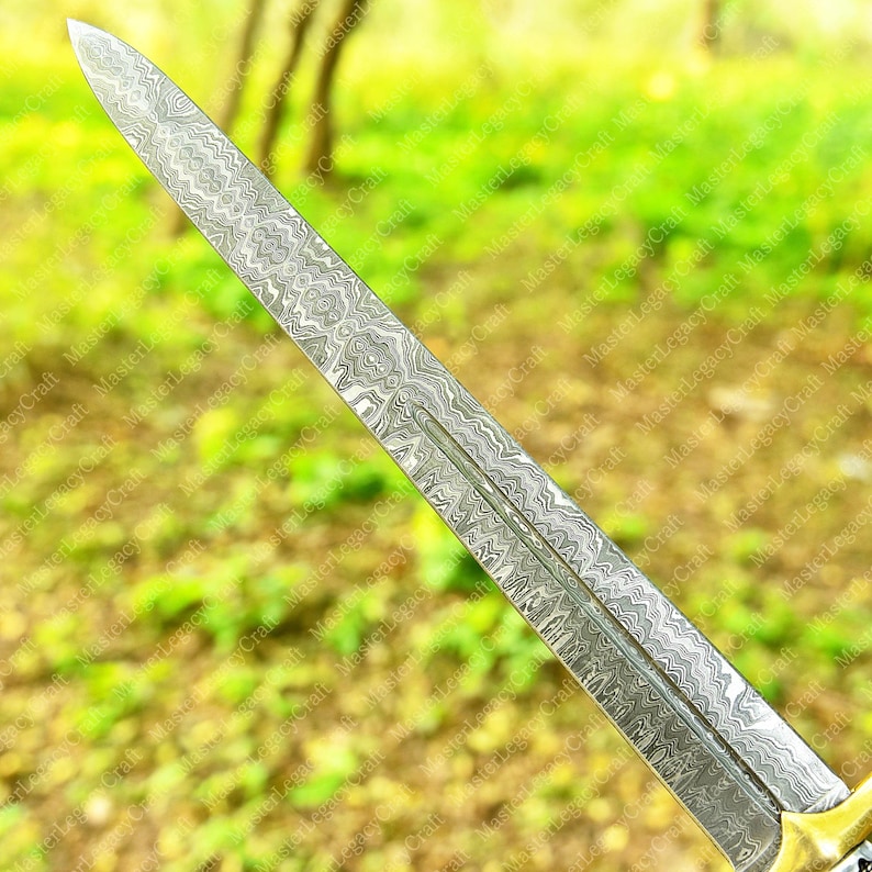 May include: Close-up of a decorative sword blade with a detailed, patterned design. The blade has a sharp point and a central groove. The handle is not visible. The blade's surface has a silver and gray color.