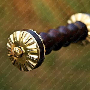 May include: Close-up of a sword hilt. The hilt features a gold-colored, sunburst-style pommel and a dark brown, wooden grip. The design suggests a historical or fantasy theme.