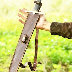 May include: A medieval-style sword with a silver blade and gold accents, sheathed in a brown leather scabbard. The hilt features a detailed design, and the scabbard has a strap for carrying. The sword is held against a blurred green background.