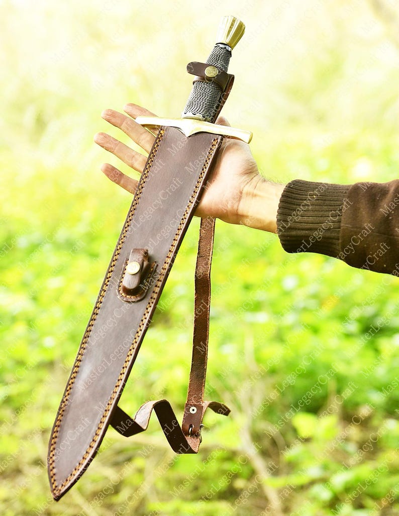 May include: A medieval-style sword with a silver blade and gold accents, sheathed in a brown leather scabbard. The hilt features a detailed design, and the scabbard has a strap for carrying. The sword is held against a blurred green background.