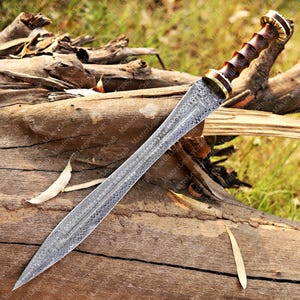 May include: A detailed view of a sword with a patterned blade and a wooden handle. The handle has a dark brown color with gold accents. The blade is silver and has a detailed pattern. The sword is lying on a piece of wood.