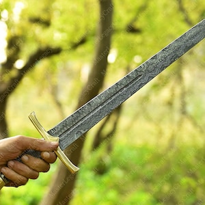 May include: A long, ornate sword with a patterned blade and a gold-colored hilt. The blade has a textured design, and the hilt features a crossguard and a decorative pommel. The sword is held against a blurred green background.
