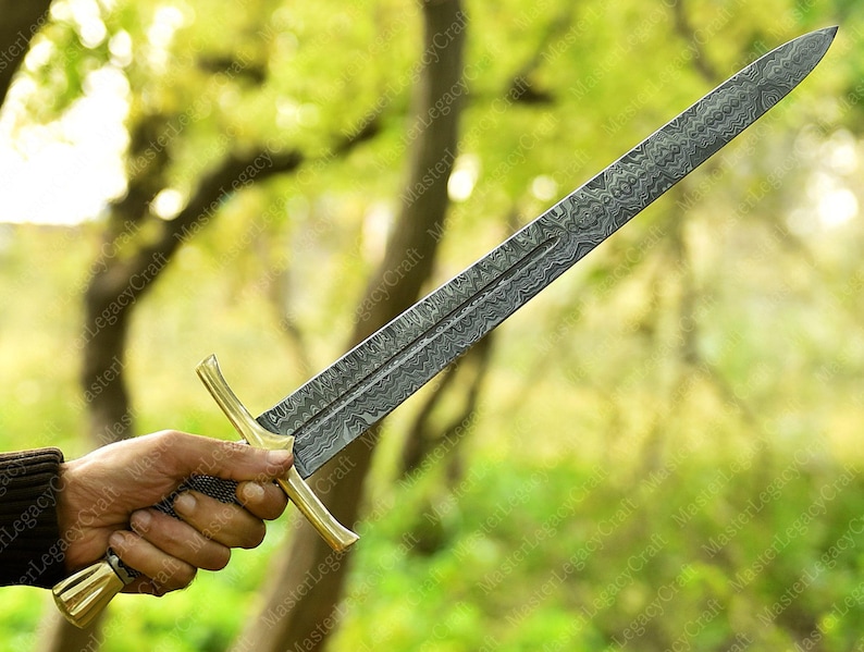 May include: A long, ornate sword with a patterned blade and a gold-colored hilt. The blade has a textured design, and the hilt features a crossguard and a decorative pommel. The sword is held against a blurred green background.