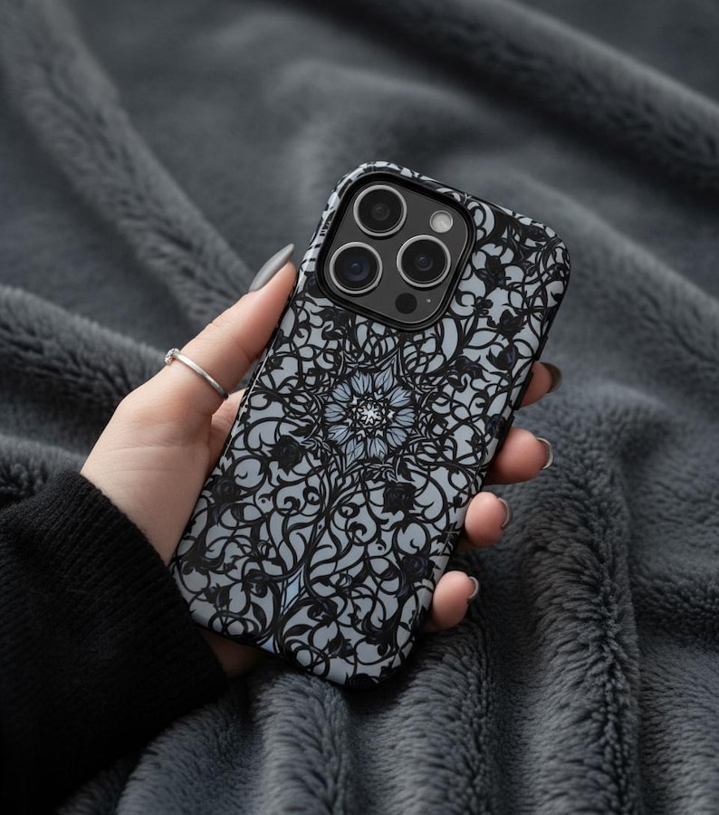 May include: A black and gray phone case with a floral mandala design. The case has a black border and a gray background with a detailed, swirling pattern. The phone case is held against a gray, textured background.