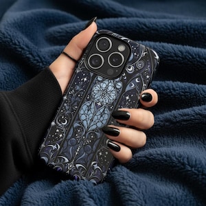 Op de afbeelding: Een zwart-blauwe telefoonhoes met een glas-in-lood ontwerp met manen, sterren en bloemelementen. De hoes heeft een donkere achtergrond met lichtblauwe en paarse accenten. De telefoonhoes wordt tegen een blauwe, getextureerde achtergrond gehouden.