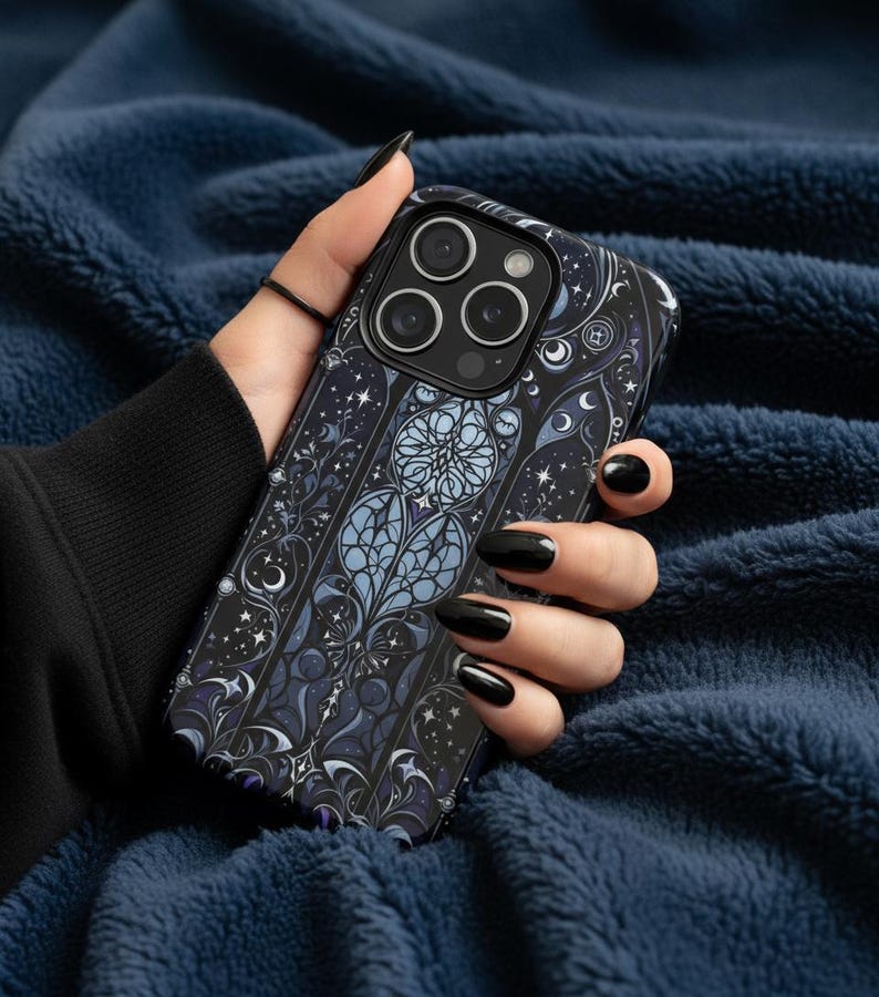 Op de afbeelding: Een zwart-blauwe telefoonhoes met een glas-in-lood ontwerp met manen, sterren en bloemelementen. De hoes heeft een donkere achtergrond met lichtblauwe en paarse accenten. De telefoonhoes wordt tegen een blauwe, getextureerde achtergrond gehouden.