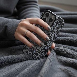 May include: A black and gray phone case with a floral design is held in a hand. The case has a matte finish and a raised camera bump. The background is a gray, plush blanket. The person is wearing a gray sweater and a silver ring.