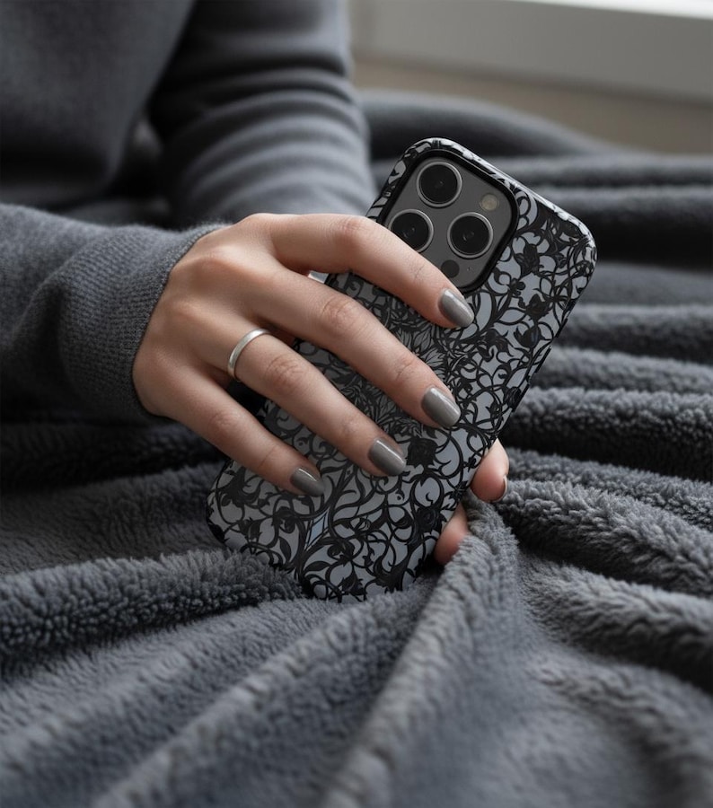 May include: A black and gray phone case with a floral design is held in a hand. The case has a matte finish and a raised camera bump. The background is a gray, plush blanket. The person is wearing a gray sweater and a silver ring.
