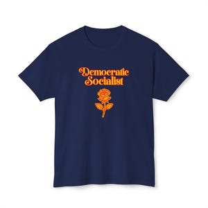 May include: A navy blue short-sleeve t-shirt featuring the text "Democratic Socialist" in a bold orange font. Below the text is a graphic illustration of a single rose in the same orange colour, centred on the chest of the garment.