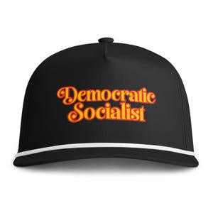 May include: A black baseball cap featuring the text Democratic Socialist embroidered in a bold orange and red font across the front. The cap includes a white cord detail running horizontally across the base of the crown above the brim.