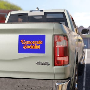 May include: A rectangular sticker applied to the tailgate of a light-coloured pickup truck. The sticker features a solid blue background with the text "Democratic Socialist" printed in a bold, orange, retro-style serif font centred on the surface.