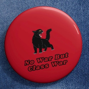 Op de afbeelding: Een rode ronde button met een zwarte kat illustratie met open mond en de tekst "No War But Class War". De button heeft een glanzende afwerking en staat op een denim achtergrond.