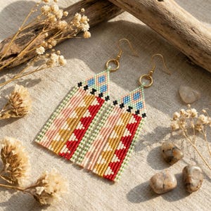 May include: A pair of handmade macrame earrings with a geometric design. The earrings feature red, pink, gold, and blue patterns, and gold-coloured ear wires. The earrings are displayed on a neutral linen surface.