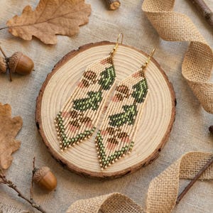 May include: A pair of beaded earrings with a fringe design. The earrings feature a pattern of green, pink, and brown beads on a cream-colored background. They are displayed on a wooden slice with acorns and leaves.