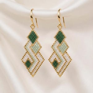 Op de afbeelding: Een paar kralen oorbellen met een geometrisch ontwerp. De oorbellen hebben goudkleurige haken en zijn gemaakt met groene, witte en gouden kralen. Het ontwerp omvat ruitvormen en een herhalend patroon.