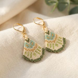 May include: A pair of beaded earrings with gold-colored hooks. The earrings feature a teardrop shape with a pattern of light blue, green, and gold beads. The earrings are on a white surface.