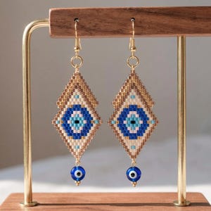 May include: A pair of beaded earrings with a diamond shape, featuring a blue and white evil eye design. The earrings are gold-colored and have a small blue bead at the bottom. They are displayed on a wooden stand.
