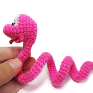 May include: A pink crocheted snake toy with white eyes and a red tongue. The snake is curled up in a coil.