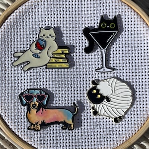 May include: Four enamel pins on a white background. The pins feature a white cat with a wine glass, a black cat in a martini glass, a dachshund with heart-shaped sunglasses, and a sheep made of yarn.