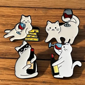 May include: Four white cat enamel pins with black outlines. Each cat is depicted with a wine theme: one reclining with a glass, one holding a glass, one wearing sunglasses with a bottle, and one drinking from a glass.