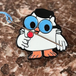 May include: A white enamel pin shaped like an owl wearing a black beret and large blue glasses, holding a red lollipop in one wing. A silver sewing needle with a light blue thread is positioned diagonally across the front of the owl pin.