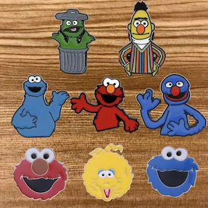 May include: A collection of colorful character pins featuring Sesame Street characters. The pins include Oscar the Grouch, Bert, Cookie Monster, Elmo, Grover, and Big Bird. Each pin has a detailed design with vibrant colors and outlines.