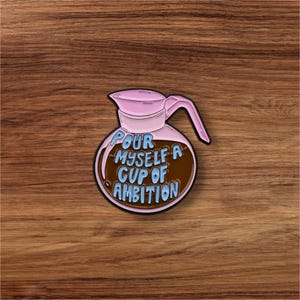 May include: A pink and brown enamel pin in the shape of a coffee pot. The pin features the text "Pour Myself A Cup Of Ambition" in blue lettering. The pin is set against a wooden background.